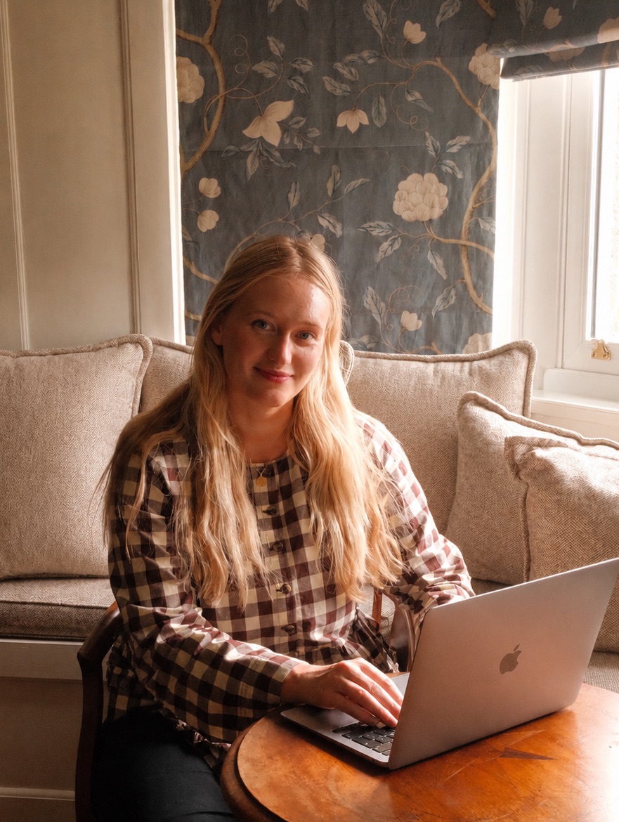 Hannah at a table with her laptop, in a sitting room with a floral blind